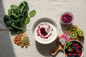 A flat lay of gut-healthy foods including yogurt, fermented vegetables, fresh fruits, leafy greens, nuts, and seeds on a neutral background.