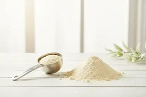 Scoop and pile of white protein powder on a white surface with green leaves in the background